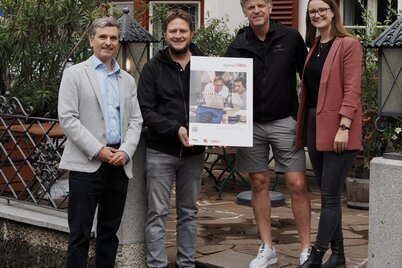 Pressefoto 2 WiR Alpbachtal Digital-Lotsen-Programm | © WiR Alpbachtal Drei Männer und eine Frau stehen vor dem Gasthof Herrnhaus in Brixlegg und halten gemeinsam eine Tafel mit der Aufschrift "Digital-Lotsen-Programm Tirol". | © WiR Alpbachtal